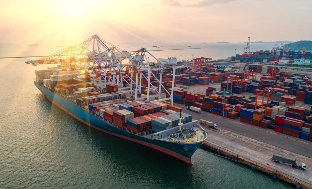 Navio cargueiro atracado em terminal portuário com movimentação de contêineres e guindastes.