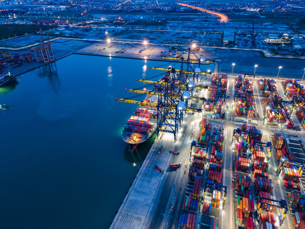 Vista aérea de terminal portuário com grande volume de contêineres e operação logística ativa.